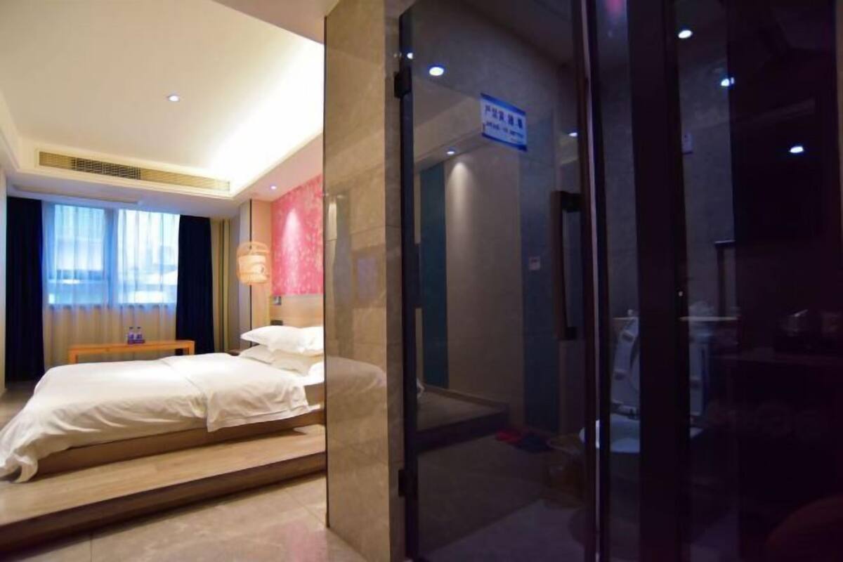 A hotel room with a bed, a bedside table, a window with curtains, and a glass door leading to a bathroom.