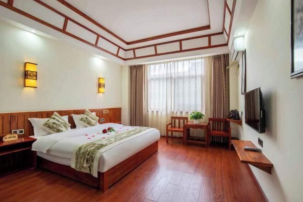 A hotel room with a wooden bed, bedside tables, a desk, a chair, a television, and a large window with curtains.