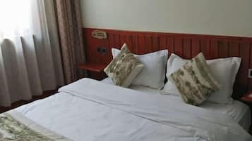 Una habitación de hotel con cama, almohadas, mesita de noche y ventana con cortinas.