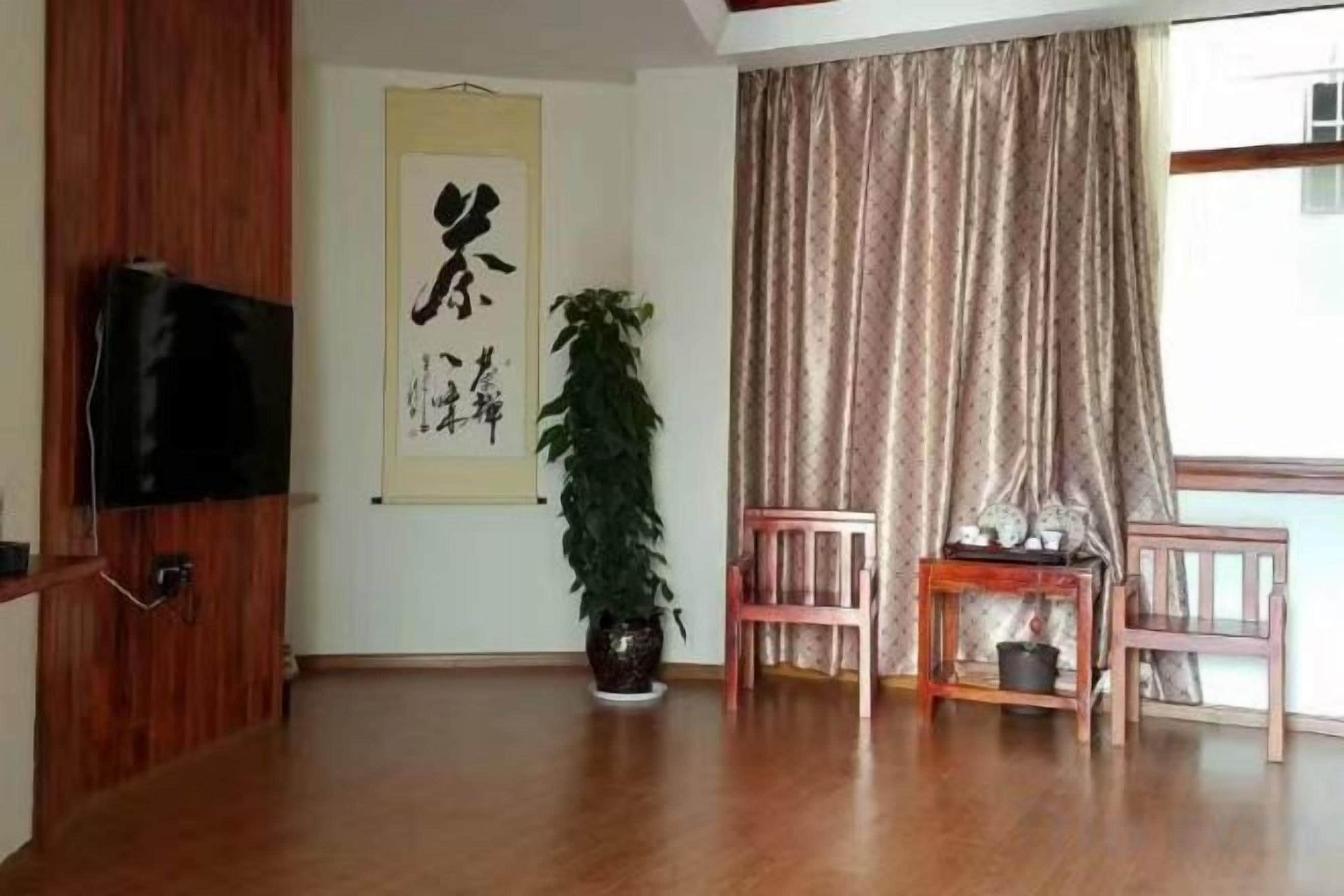 Una habitación con piso de madera, un televisor instalado en un panel de madera, una planta grande en maceta, una obra de arte de caligrafía china tradicional y una ventana con cortinas.