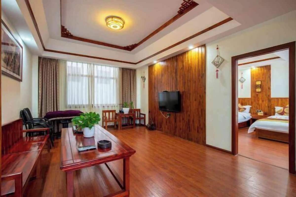 A hotel room with a wooden floor, a TV mounted on the wall, a bed with a wooden headboard, a desk with a chair, and a window with curtains.