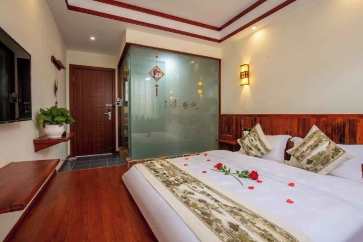 A hotel room with a bed, wooden headboard, bedside tables, a potted plant, and a sliding door.