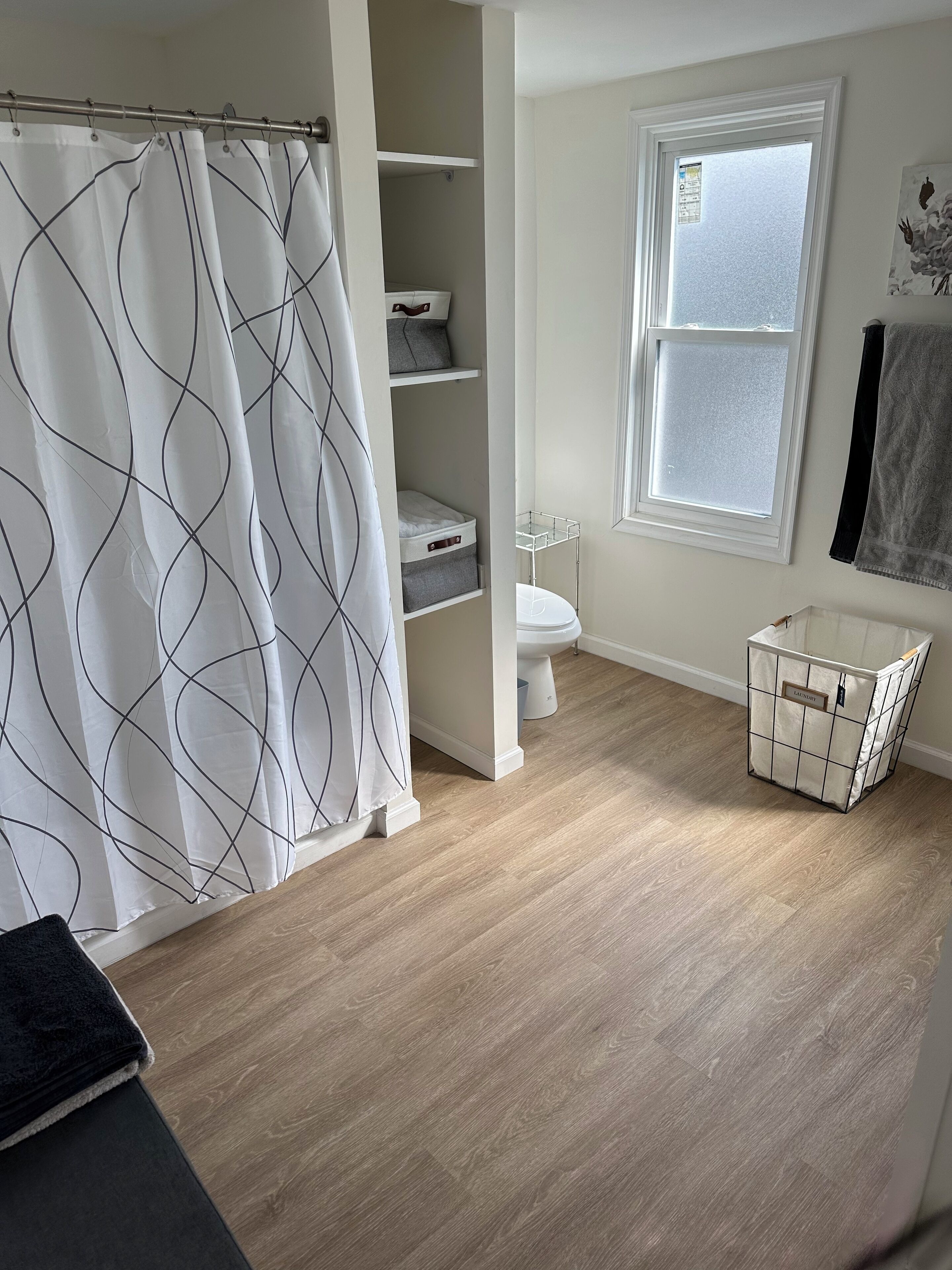 A bathroom with a shower curtain, a toilet, a window, and a laundry basket.