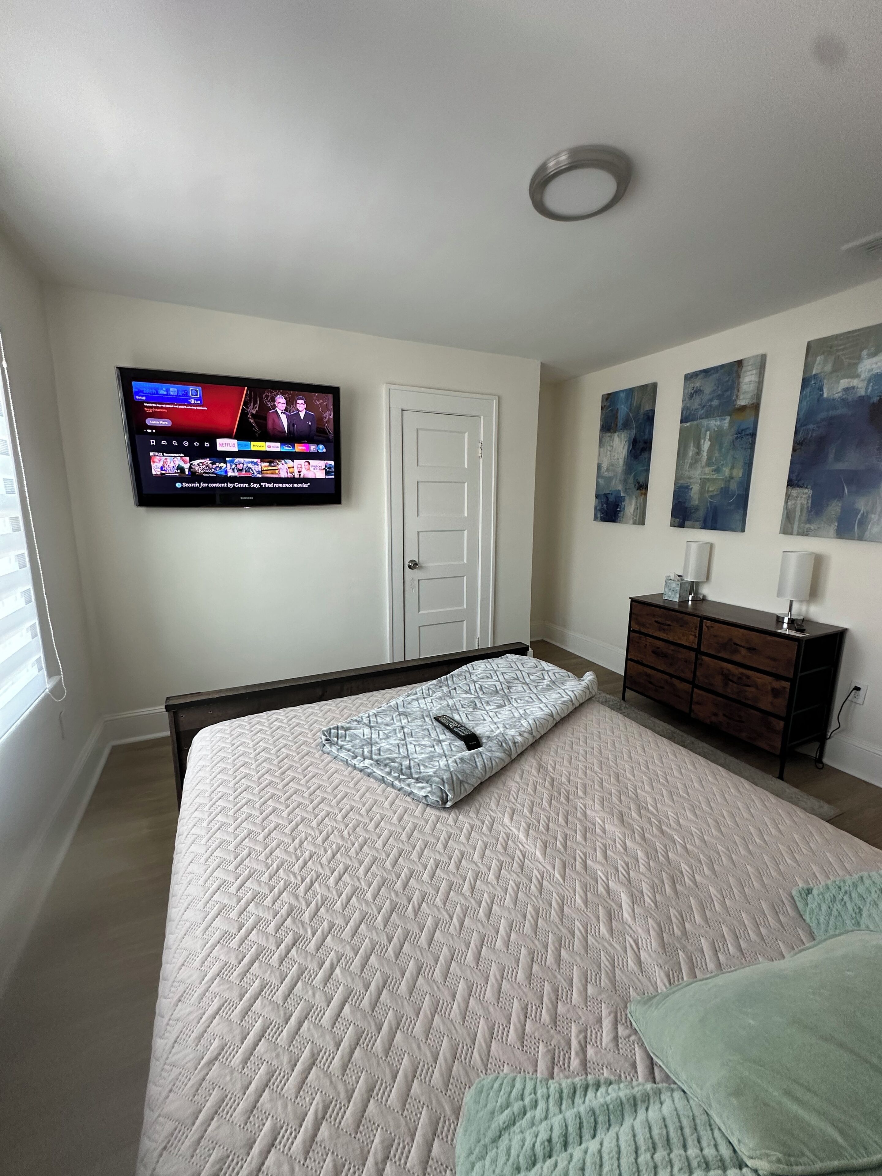 Un dormitorio con cama, televisor montado en la pared, puerta y cómoda con lámpara.