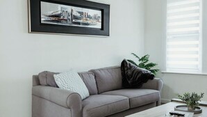 Un salon moderne avec un canapé gris, un tapis à motifs, une table basse avec des livres et une télécommande, et un tableau encadré accroché au mur.