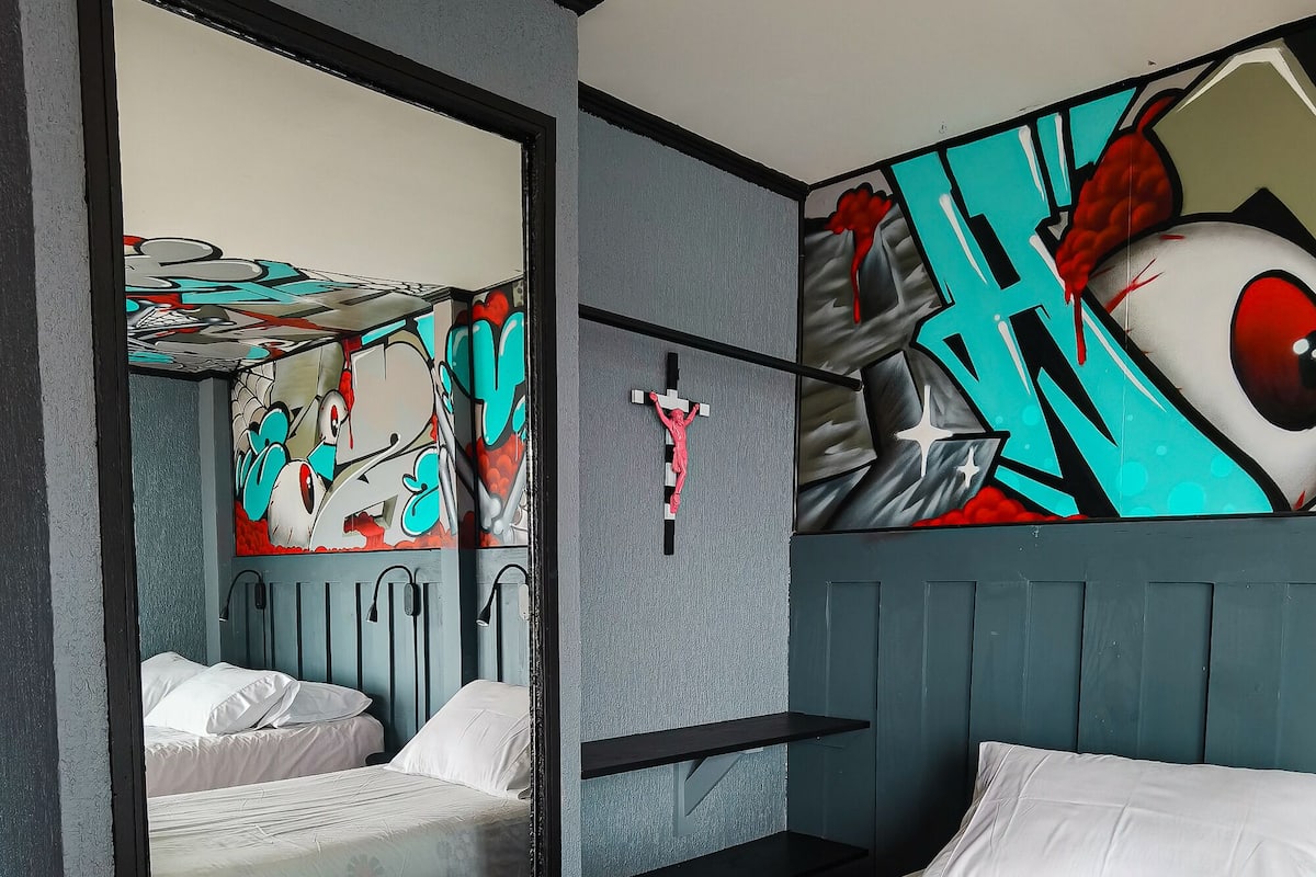 A hotel room with a bed, a mirror, a wall-mounted cross, and a colorful mural.
