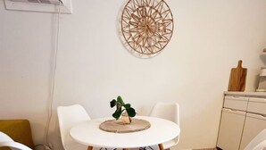 Et spiseområde med et rundt bord, fire hvide stole, en dekorativ vægdekoration, en lille plante og et køkken med skærebræt og skabe.