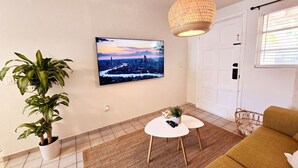 En stue med et stort fladskærms-TV monteret på væggen, en hængende vævet lampe, en potteplante, et rundt hvidt sofabord og en sennepsgul sofa.
