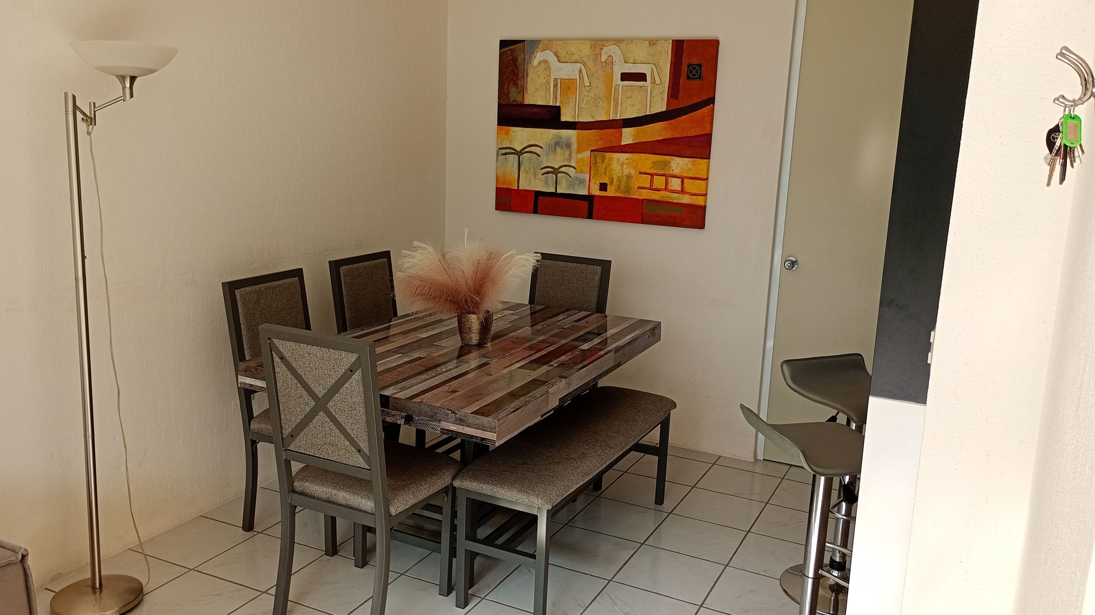A dining area with a wooden table, six chairs, a vase with dried plants, and a painting on the wall.