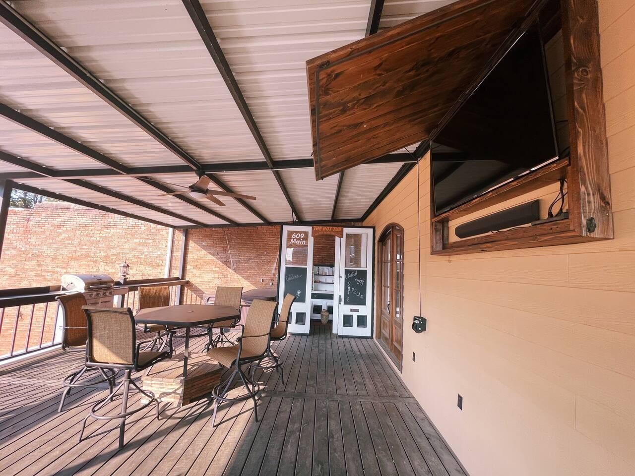 An outdoor patio with a wooden table and chairs, a grill, and a TV mounted on the wall.