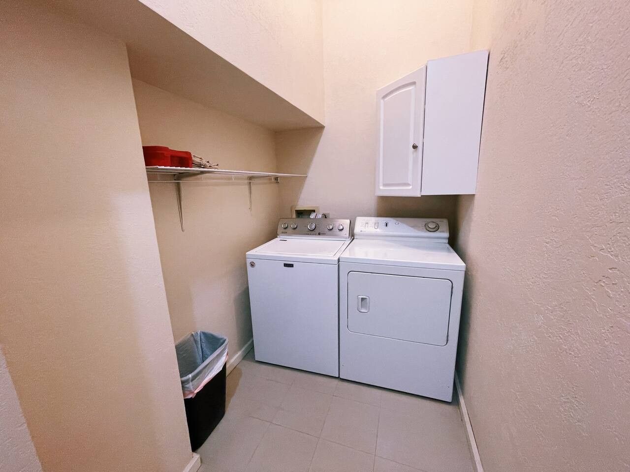 A laundry room with a washing machine and dryer, a trash bin, and a cabinet.