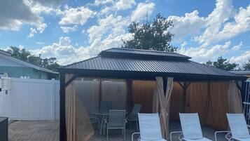A covered outdoor seating area with lounge chairs and a table, surrounded by a white fence and a wooden wall.