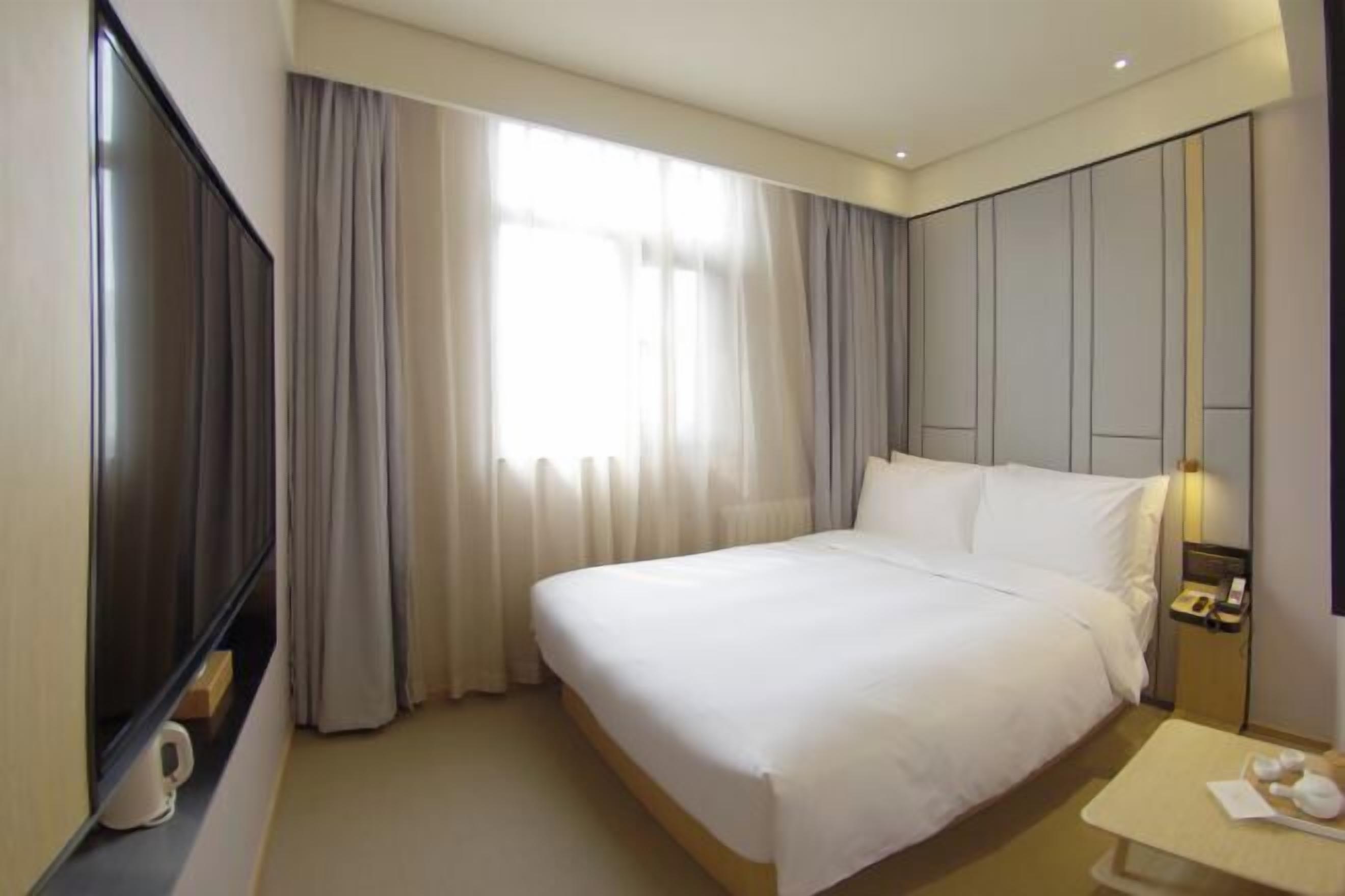 A hotel room with a large bed, a flat-screen TV, a bedside table with a lamp, and a window with curtains.