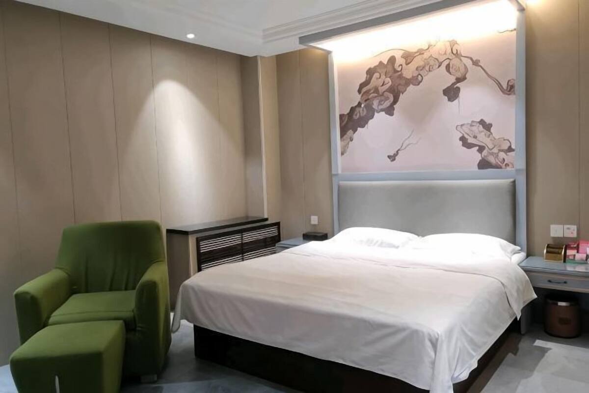 A bedroom with a bed, a green armchair, a wall-mounted artwork, and a nightstand with a lamp.