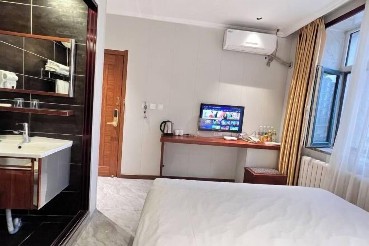A hotel room with a bed, a TV, a desk, a bathroom with a sink and mirror, and a window with curtains.