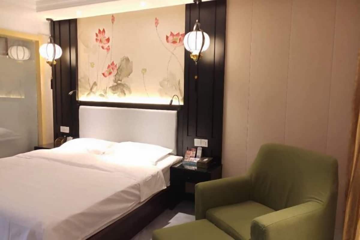 A hotel room with a bed, a green armchair, a nightstand, and a wall with a floral painting.
