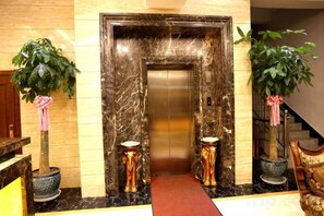 Entrada de ascensor con paredes de mármol, plantas en macetas y alfombra roja.