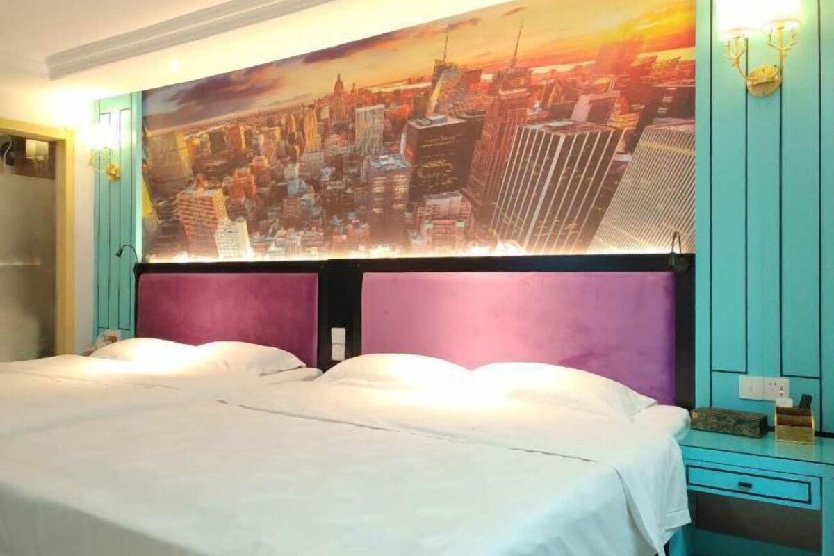 A hotel room with a large cityscape painting on the wall, a bed with white linens, and a teal-colored dresser.