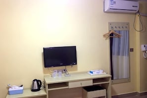 A hotel room with a desk, a television, a kettle, a cup, a tissue box, and a window with a curtain.