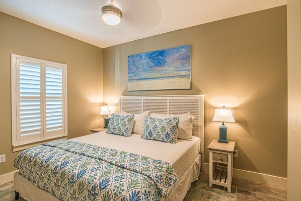 A bedroom with a bed, two bedside lamps, a painting of a beach, and a window with blinds.