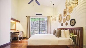 Ein Schlafzimmer mit einem hölzernen Bett, einem Schreibtisch, einem Stuhl, einem Deckenventilator und Blick auf eine Steinmauer durch eine Glastür.
