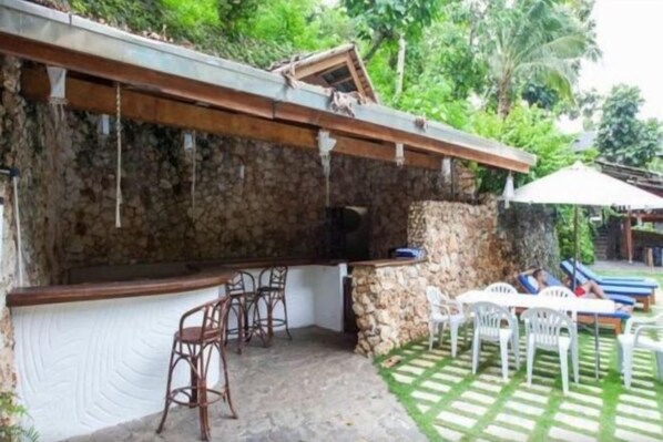 An outdoor bar area with stone walls, bar stools, and a seating area with white plastic chairs.