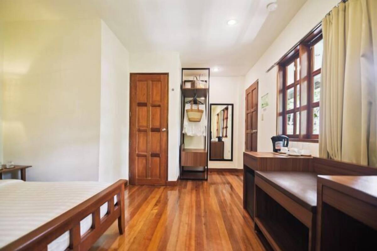 Quarto com piso de madeira, uma cama, uma escrivaninha, uma janela com cortinas e uma porta.