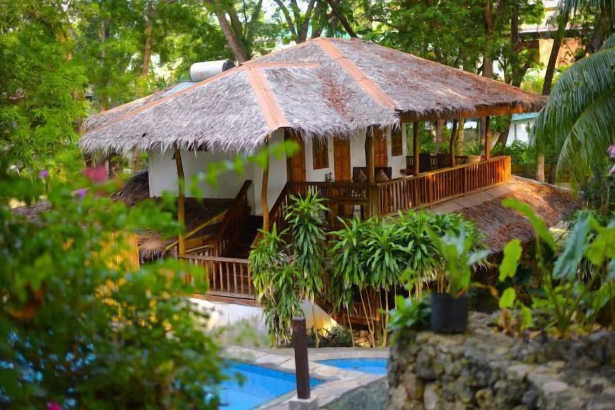 Uma construção com telhado de palha e deck de madeira, rodeada por vegetação exuberante e uma pequena piscina.