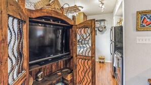 Una cocina con un televisor de pantalla plana grande, armarios de madera, un taburete pequeño y diversos electrodomésticos.