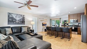 A modern living room with a sofa, a ceiling fan, and a kitchen area with a bar counter. - Screened-in Porch | Walkable to SoCO (Austin)