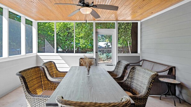 A covered patio with a dining table and wicker chairs, large windows, and a ceiling fan. - Screened-in Porch | Walkable to SoCO (Austin)