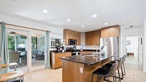 A modern kitchen with a central island, bar stools, and a dining area with a table and chairs. - Screened-in Porch | Walkable to SoCO (Austin)