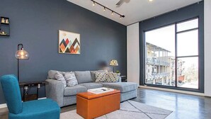 A modern living room with a grey sofa, a blue armchair, a coffee table, a lamp, and a large window with a view of buildings.