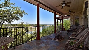A covered patio with stone flooring, wicker furniture, and a view of a river. - Lakefront Escape | PingPong + Fire Pit | Sleeps 12 (Spicewood)