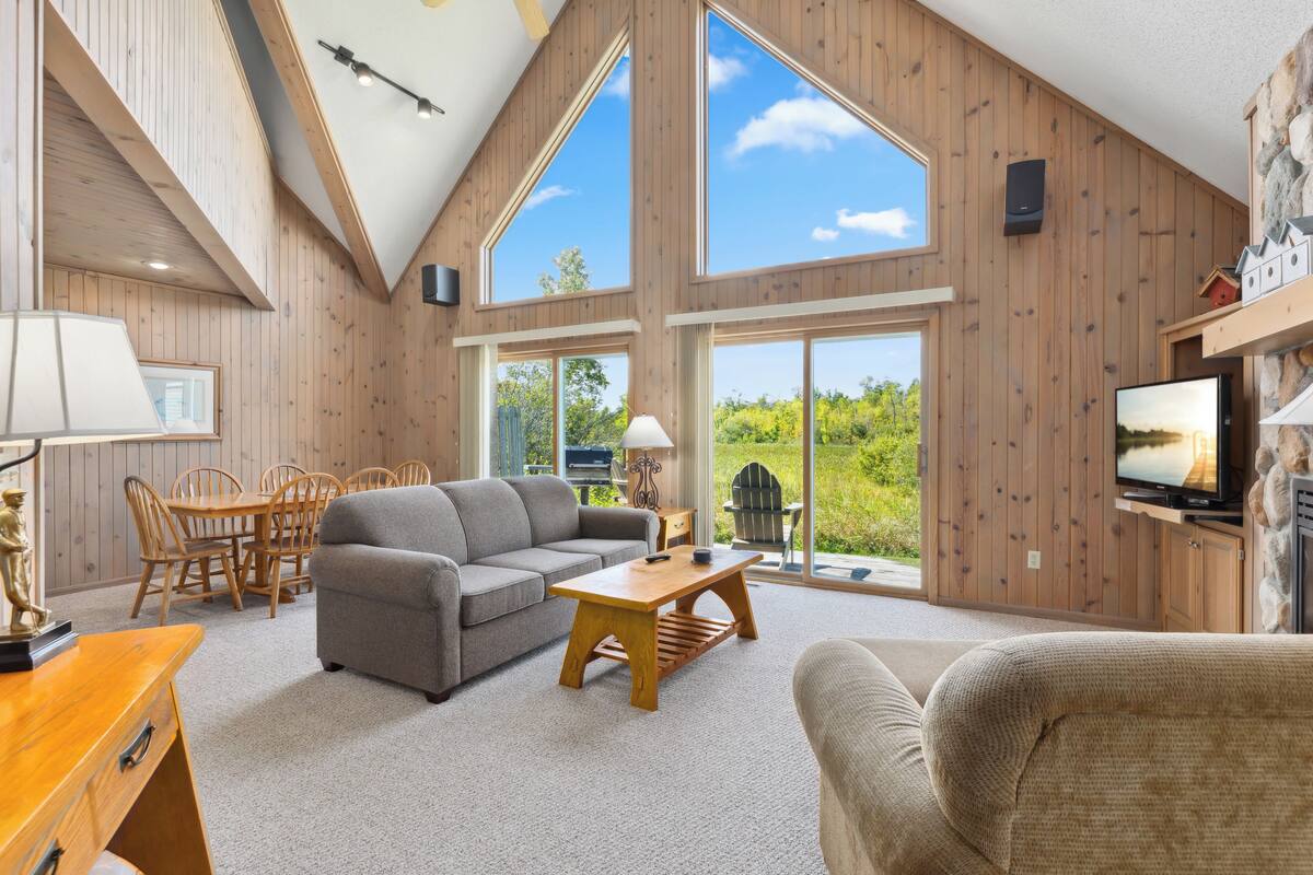 A spacious living room with a high ceiling, wooden walls, a gray sofa, a wooden coffee table, and a television.