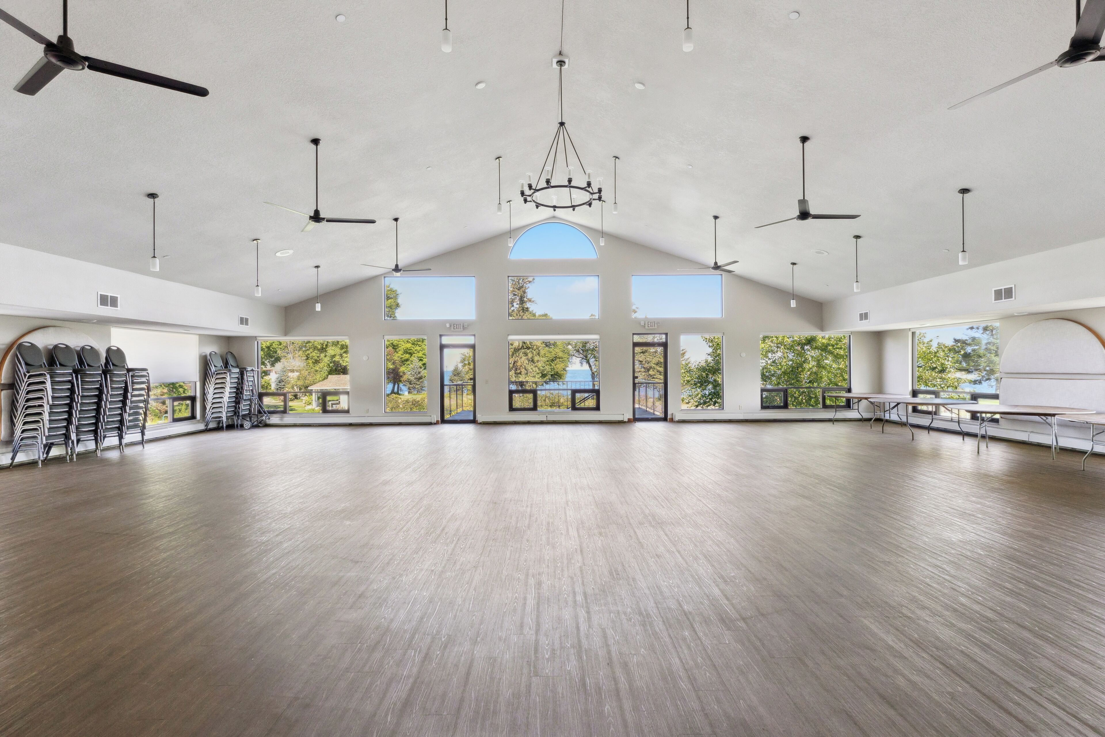 A spacious room with a high ceiling, multiple ceiling fans, a chandelier, and large windows offering views of trees and a body of water.