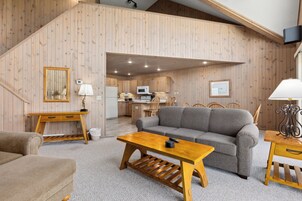 Una sala acogedora con un sofá, una mesa de centro de madera y un televisor. Se ve una cocina al fondo con armarios de madera y un microondas.
