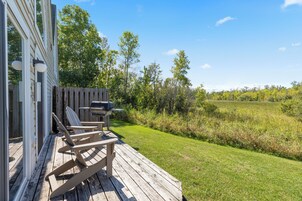 Una terraza de madera con sillas Adirondack y una parrilla, con vistas a un campo cubierto de césped y árboles.