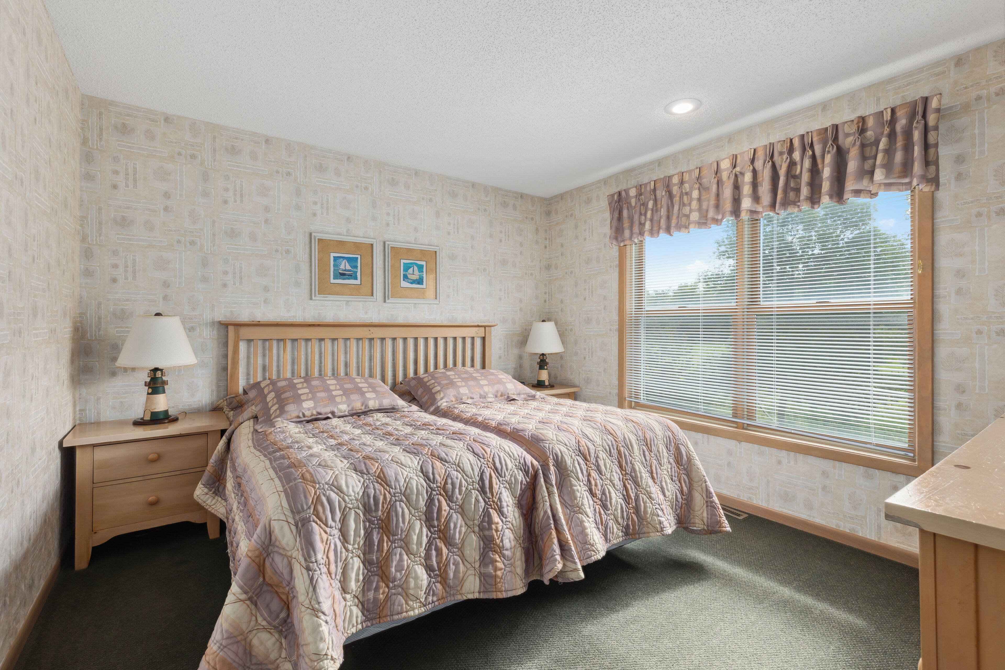 A bedroom with a bed, two bedside tables with lamps, a window with blinds, and patterned wallpaper.