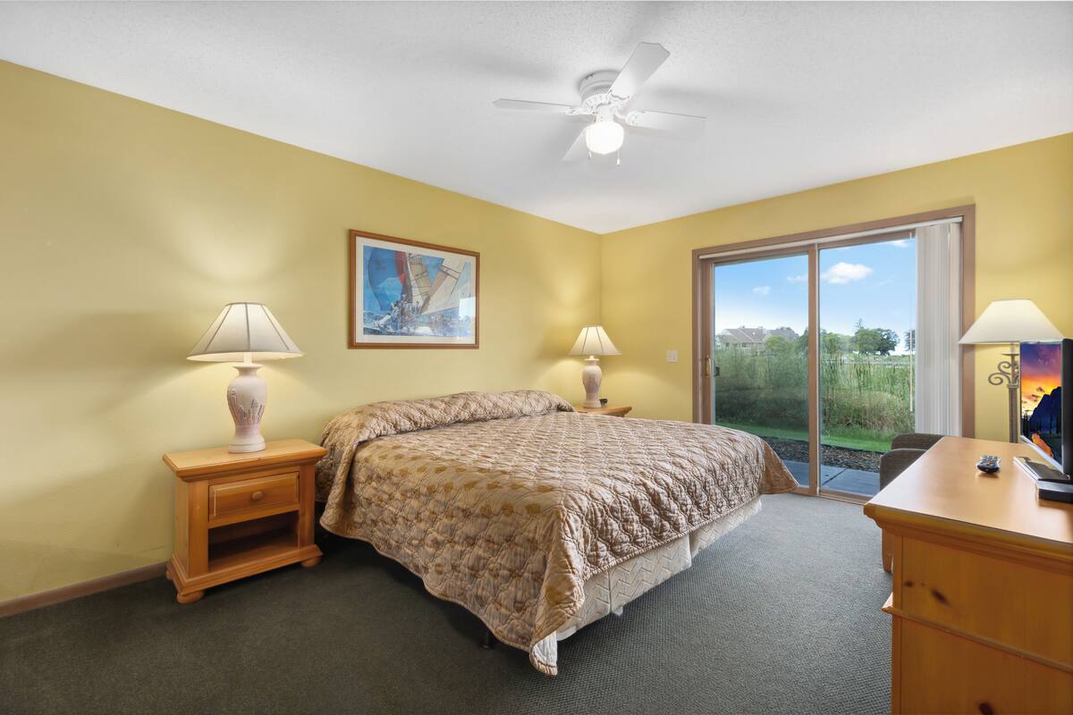 A bedroom with a bed, two bedside tables with lamps, a painting on the wall, and a view through a large window.