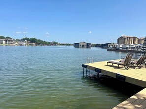 Un muelle frente al agua con sillones de descanso, piscina y edificios al fondo.