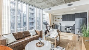 Een moderne woonkamer met een bank, fauteuil en salontafel, een keuken met donkere kastjes en grote ramen met uitzicht op de stad.