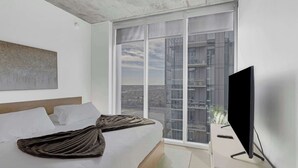 Een moderne hotelkamer met een groot bed, een flatscreen-tv en een uitzicht op een stadsbeeld door middel van kamerhoge ramen.
