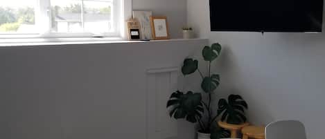 A modern living room with a large window, a flat-screen TV mounted on the wall, and a wooden dining table with two chairs.