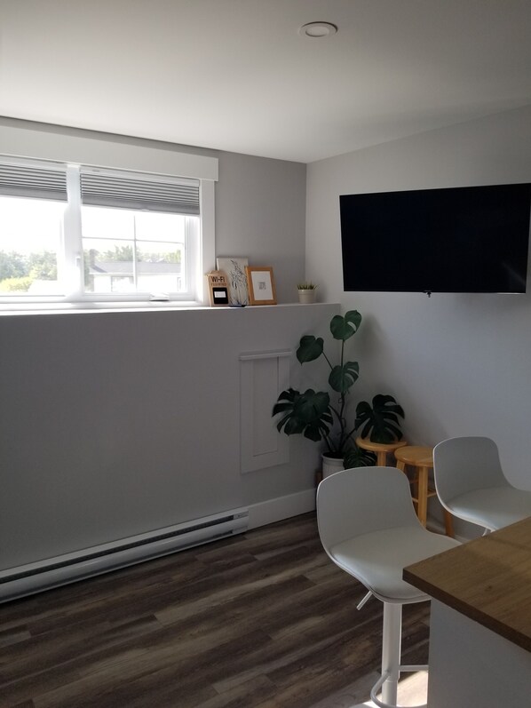 A modern living room with a large window, a flat-screen TV mounted on the wall, and a wooden dining table with two chairs.
