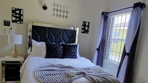 Een slaapkamer met een bed, een plafondventilator, een nachtkastje, een lamp, een raam met gordijnen en wanddecoratie.