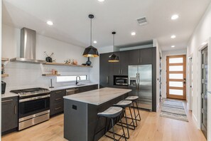 A modern kitchen with a central island, stainless steel appliances, pendant lighting, and a wooden floor.