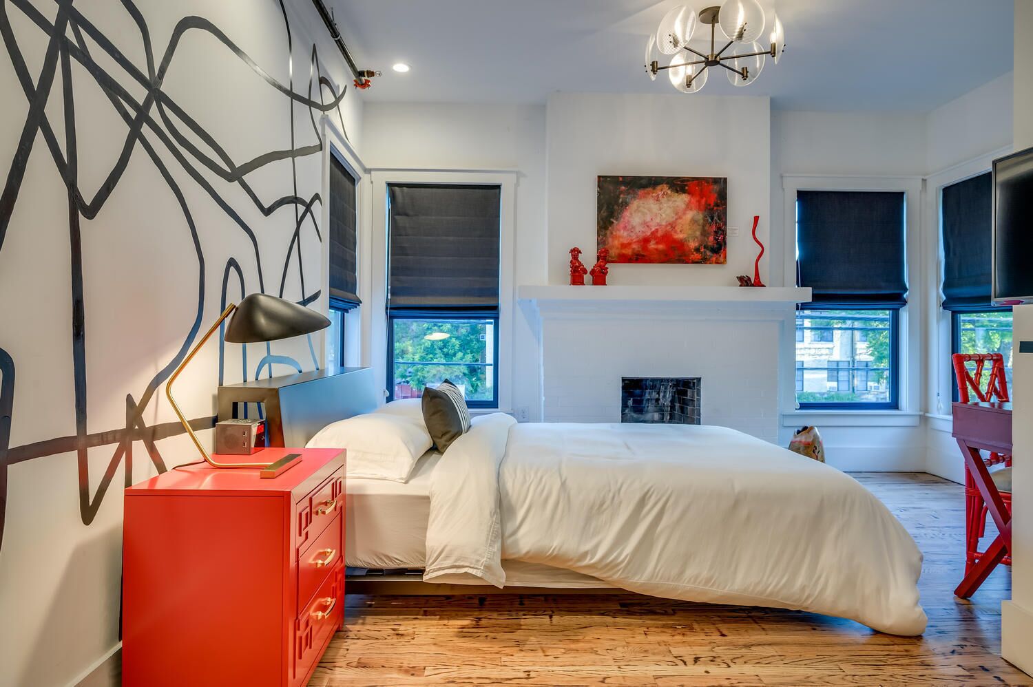A modern bedroom with a bed, a red dresser, a fireplace, and abstract wall art.