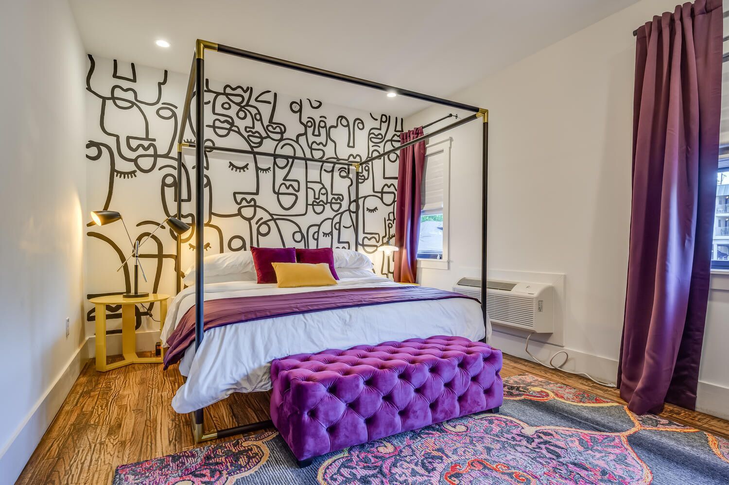 A four-poster bed with a unique wall mural, a purple ottoman, and a patterned rug.