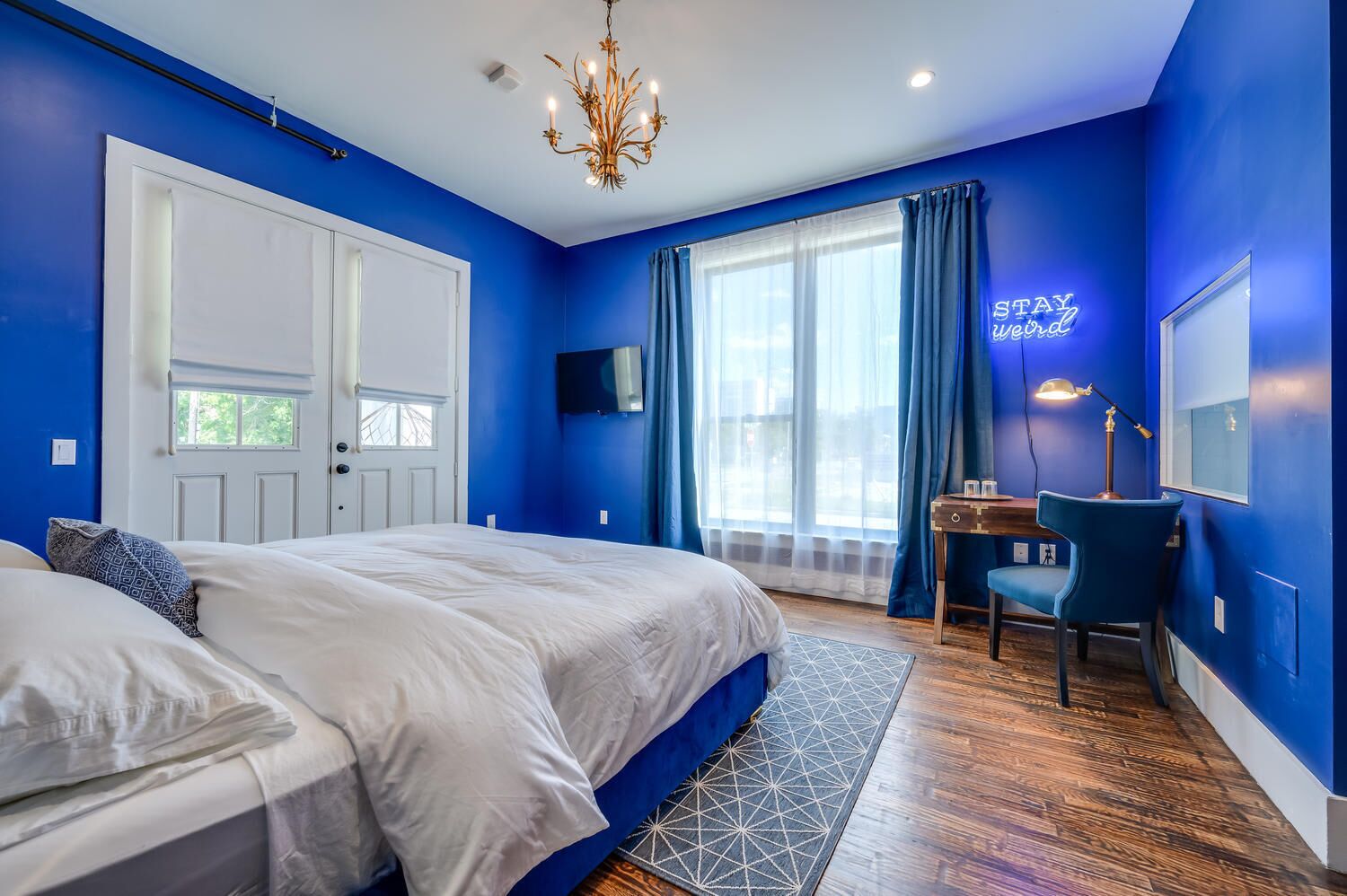 A bedroom with a large bed, a desk, a chair, a TV, and a chandelier.